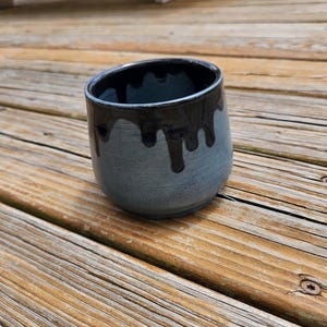 May include: A ceramic cup with a rounded shape, featuring a textured matte grey finish on the lower portion and a glossy black glaze dripping from the rim down the sides. The interior of the cup is finished in a smooth, solid black glaze.
