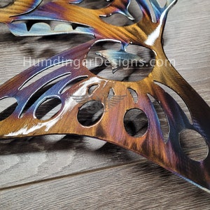 May include: A metal butterfly wall art with a cut-out design and a rainbow iridescent finish. The butterfly is on a wooden surface. The text "HumdingerDesigns.com" is visible in the image.