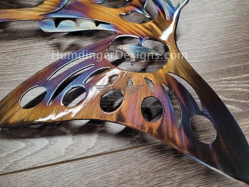 May include: A metal butterfly wall art with a cut-out design and a rainbow iridescent finish. The butterfly is on a wooden surface. The text "HumdingerDesigns.com" is visible in the image.