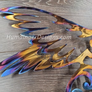 May include: A metal wall art sculpture of a bird with wings spread wide. The sculpture is made of steel and has a rainbow iridescent finish. The sculpture is mounted on a wooden surface.