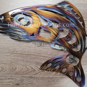 May include: A metal sculpture of a salmon with a rainbow iridescent finish. The sculpture is cut out and has a detailed design. The sculpture is on a wooden surface.