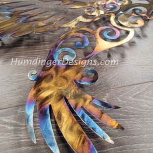 May include: A metal wall art sculpture of a stylized bird with intricate details. The bird's wings are spread wide and have a blue and gold iridescent finish. The sculpture is made of metal and has a rustic, hand-crafted look.