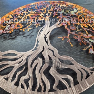 K&ouml;nnte beinhalten: Eine runde Wandkunst aus Metall in Form eines Lebensbaums. Der Baum hat ein detailliertes, filigranes Design mit einer Kupfer-Oberfl&auml;che. Die &Auml;ste sind in einem warmen Orangeton gehalten, w&auml;hrend die Wurzeln silbern sind.