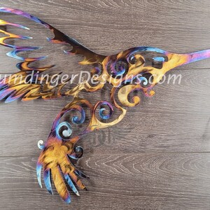 May include: A colorful metal hummingbird wall art with intricate details and a swirling design. The hummingbird is in flight with its wings spread out. The metal is a mix of blue, purple, orange, and yellow.