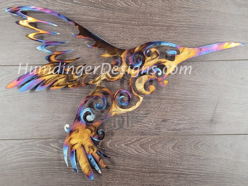 May include: A colorful metal hummingbird wall art with intricate details and a swirling design. The hummingbird is in flight with its wings spread out. The metal is a mix of blue, purple, orange, and yellow.