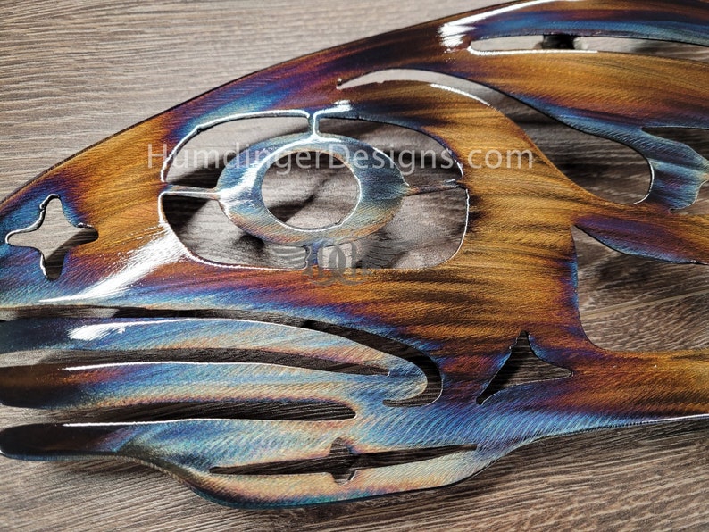May include: A metal wall art piece in the shape of a fish with a blue, purple, and brown iridescent finish. The fish has a cut-out design and the text "HumdingerDesigns.com" is visible on the surface.