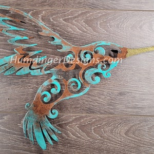 May include: A turquoise and brown metal hummingbird wall art with a gold beak. The hummingbird is in flight with its wings spread out. The metal has a distressed finish, giving it a rustic look.