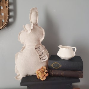 May include: A white fabric bunny with a tag that says "Hop" sitting on a black surface with a white creamer and two books stacked on top of each other.