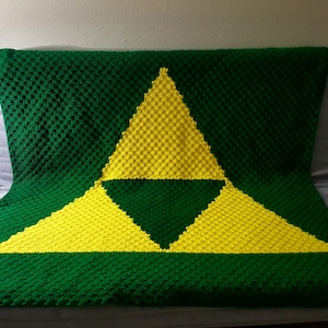 May include: A green and yellow crocheted blanket with a triangular pattern, resembling the Triforce from the Legend of Zelda video game series.