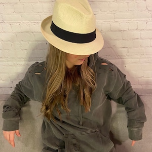 May include: A person wearing a dark gray distressed hoodie with a white fedora hat with a black band.