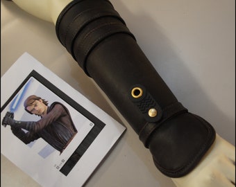 May include: Dark brown leather arm bracer with gold-colored rivets and a decorative strap. The bracer is shown on a mannequin arm next to a printed image of a character holding a lightsaber.