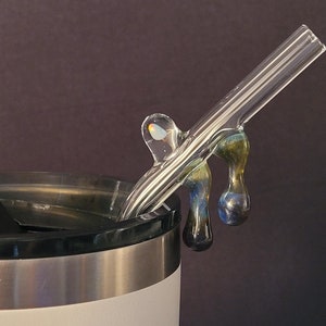 May include: A clear glass straw with a decorative, colorful glass accent. The straw is angled over the rim of a white and silver tumbler. The accent features blue, green, and gold hues.