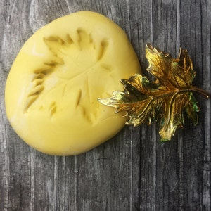 May include: A yellow silicone mold shaped like a maple leaf, with a gold-colored metal maple leaf on a wooden surface.