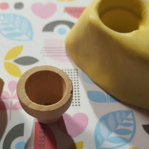May include: Two small, light brown, ceramic flower pots, one with a slightly wider opening than the other, are sitting on a colorful patterned surface. A yellow silicone mold with two circular cavities is also on the surface.