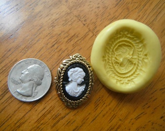 May include: A silicone mold for making cameo jewelry. The mold is yellow and has a raised design of a woman's profile in an oval frame. A quarter is shown for size comparison.