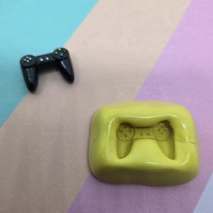 May include: A black miniature video game controller next to a yellow silicone mold of a controller. The controller has colored buttons and a sleek design. The mold is a bright yellow color.