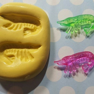 May include: A yellow silicone mold with two shrimp shapes. Two plastic shrimp, one green and one pink, are shown next to the mold.