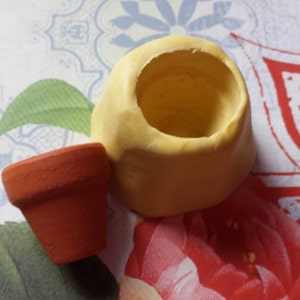 May include: A small, yellow ceramic planter with a round opening at the top. A smaller, terracotta planter sits next to it.