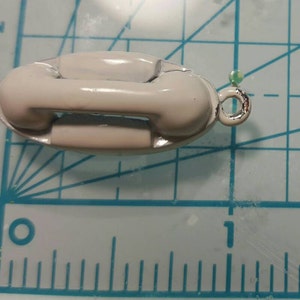 May include: A white ceramic telephone charm with a silver loop. The charm is approximately 2.5 cm long and 1.25 cm wide.