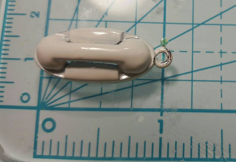May include: A white ceramic telephone charm with a silver loop. The charm is approximately 2.5 cm long and 1.25 cm wide.