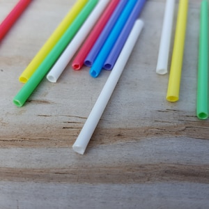 May include: A collection of colorful plastic straws in red, yellow, green, blue, and white, scattered on a light brown wooden surface. A single wooden stick is also visible. The straws are cylindrical and appear to be new.