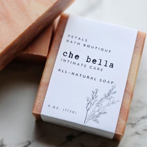 May include: A close-up of a bar of all-natural soap with a white label. The label reads "che bella INTIMATE CARE" and "PETALS BATH BOUTIQUE". The soap is 4 oz. (113g). The soap has a light brown color.