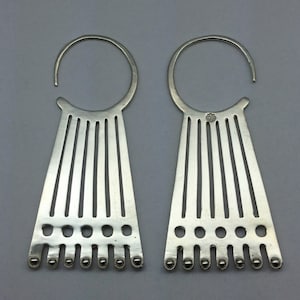 May include: A pair of silver-toned earrings. Each earring features a large circular top with a descending, fan-shaped design. The design includes vertical lines and small, round accents at the bottom. The earrings are displayed against a gray background.