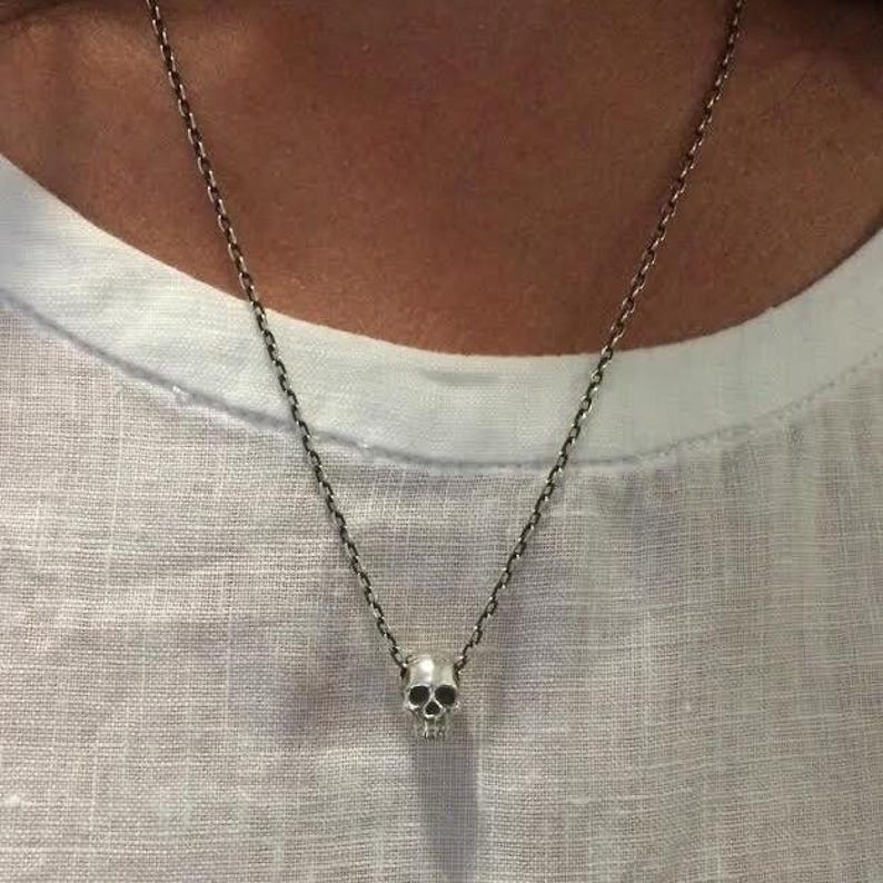 May include: A silver-toned necklace featuring a small, detailed skull pendant suspended from a delicate, textured chain. The metal has a polished finish with darkened recessed areas that highlight the anatomical features of the skull design.