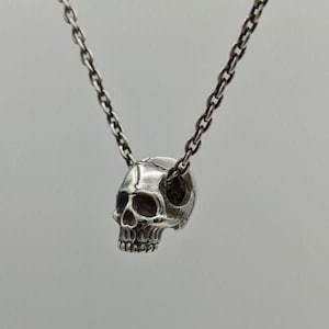 May include: A silver-toned metal necklace featuring a small, detailed skull pendant suspended from a delicate link chain. The skull is rendered with realistic anatomical features, including defined eye sockets, a nasal cavity, and teeth.
