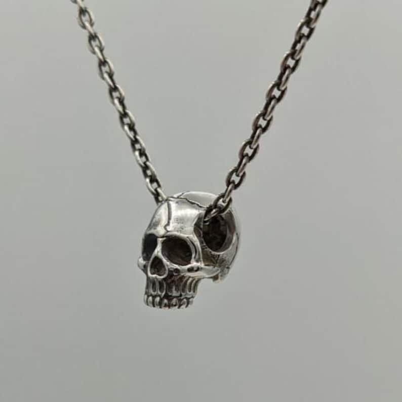 May include: A silver-toned metal necklace featuring a small, detailed skull pendant suspended from a delicate link chain. The skull is rendered with realistic anatomical features, including defined eye sockets, a nasal cavity, and teeth.