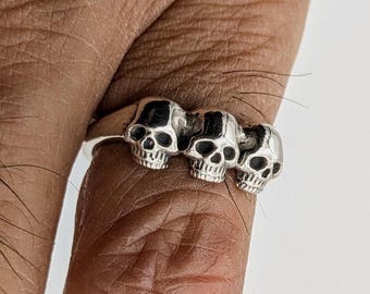 May include: A silver-toned ring featuring three distinct skull motifs arranged in a row along the band. The skulls are rendered with dark, recessed detailing in the eye sockets and nasal cavities, creating a high-contrast look on the metallic surface.