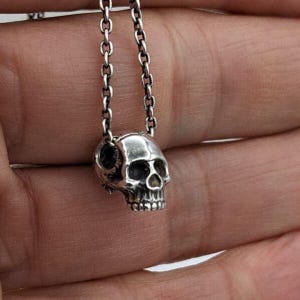 May include: A silver-toned metal skull pendant suspended from a matching silver-toned metal chain. The skull features detailed eye sockets, a nasal cavity, and defined teeth, with the chain passing through the top of the skull structure.