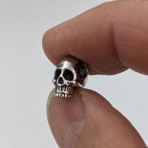 May include: A small, metallic silver-toned bead shaped like a human skull. The bead features detailed, recessed dark areas around the eye sockets, nose cavity, and teeth, providing a high-contrast, three-dimensional appearance to the skull design.