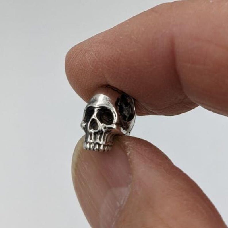 May include: A small, metallic silver-toned bead shaped like a human skull. The bead features detailed, recessed dark areas around the eye sockets, nose cavity, and teeth, providing a high-contrast, three-dimensional appearance to the skull design.