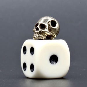 May include: A metallic silver-toned skull bead resting on top of a white six-sided die. The die features black circular pips, with four pips visible on the front face and one pip visible on the side face. The skull has detailed eye sockets.