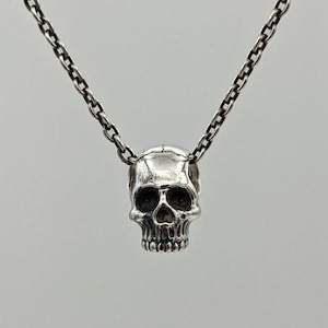May include: A silver-toned metal necklace featuring a detailed skull pendant suspended from a matching link chain. The skull has deep-set eye sockets, a defined nasal cavity, and visible teeth, with a polished finish that reflects light across its surface.
