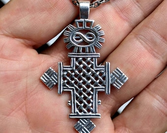 May include: A silver pendant shaped like a cross with an intricate woven pattern throughout the center. The top features a sunburst design surrounding an infinity symbol. The cross has square-shaped extensions on the sides and bottom, all suspended from a silver chain.