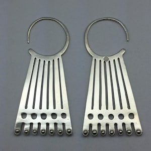 May include: A pair of silver-toned earrings featuring a circular top hook connected to a fan-shaped pendant. The pendant consists of six vertical slats, a horizontal row of six circular cutouts, and a bottom edge with six small spherical beads.