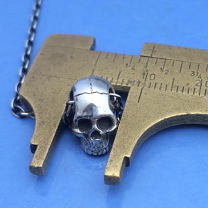 May include: A silver-colored metal skull bead attached to a thin chain, positioned between the jaws of a brass caliper. The caliper shows the skull bead measures approximately 0.5 inches in width. The skull features detailed eye sockets.