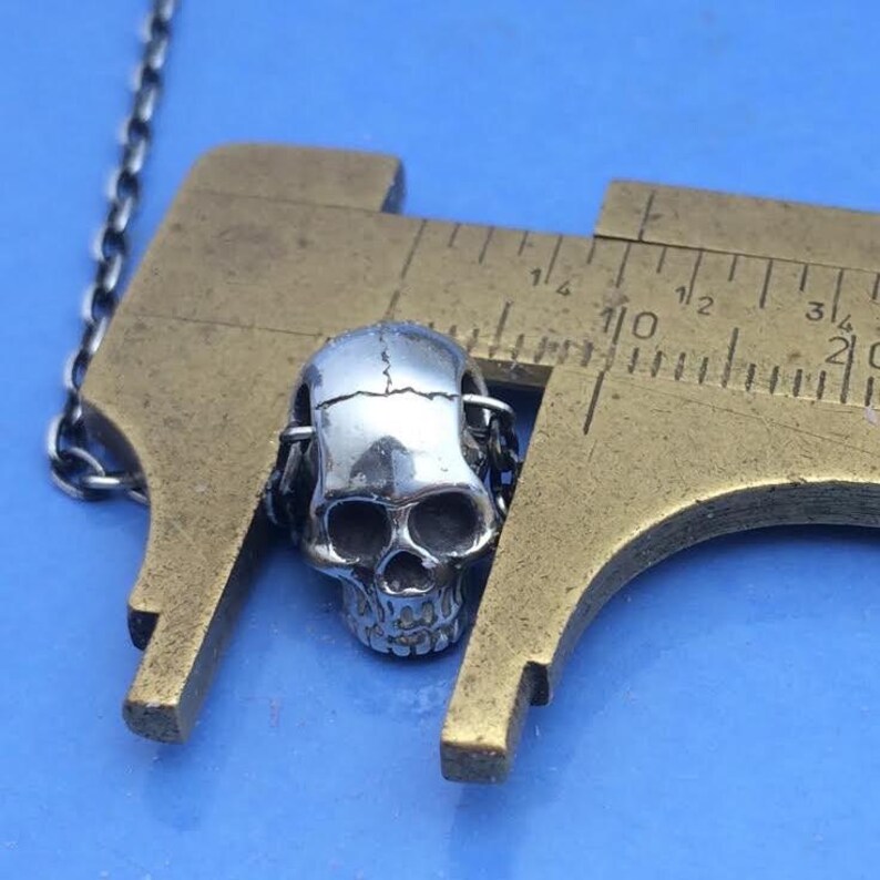 May include: A silver-colored metal skull bead attached to a thin chain, positioned between the jaws of a brass caliper. The caliper shows the skull bead measures approximately 0.5 inches in width. The skull features detailed eye sockets.