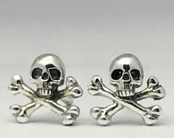 May include: A pair of silver-toned stud earrings, each featuring a detailed skull and crossbones design. The metal has a polished, reflective finish with darkened recessed areas to highlight the facial features of the skulls and the bone texture.