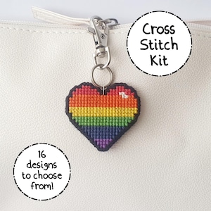 May include: A heart-shaped cross-stitch kit keychain with a rainbow design, attached to a silver clasp. The keychain is black around the edges. Text on the image reads "Cross Stitch Kit" and "16 designs to choose from!"
