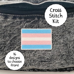 May include: A cross stitch kit featuring a transgender pride flag design. The kit is displayed on a black denim background. The text "Cross Stitch Kit" is written in a circle above the flag. The text "16 designs to choose from!" is written in a circle below the flag.