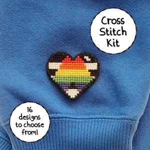 May include: A cross stitch kit featuring a heart design with rainbow colors and a black border. The kit is on a blue background. Text on the image says "Cross Stitch Kit" and "16 designs to choose from!"
