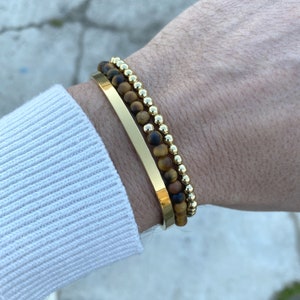 Peut inclure: Un ensemble de trois bracelets en or. Un bracelet est une simple bande d'or, et les deux autres sont des bracelets à perles avec des pierres de œil de tigre.