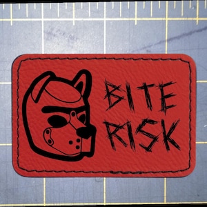 May include: A rectangular red patch with rounded corners and black stitched edges. The patch features a black graphic of a puppy hood mask on the left and the text BITE RISK in a distressed black font on the right, set against a grid mat.