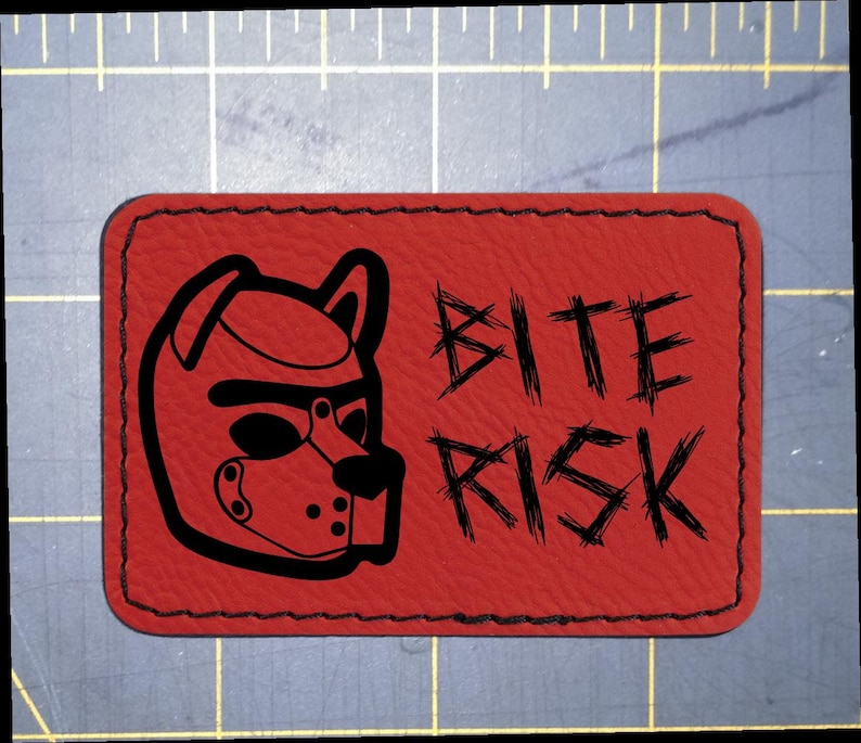 May include: A rectangular red patch with rounded corners and black stitched edges. The patch features a black graphic of a puppy hood mask on the left and the text BITE RISK in a distressed black font on the right, set against a grid mat.