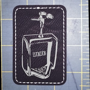 May include: A rectangular black leather patch with rounded corners and white perimeter stitching. The center features a white graphic illustration of a wall-mounted urinal with the word HERE printed in a block font on the front surface.
