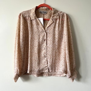 May include: A vintage button-down blouse with a beige and brown swirl pattern. The long-sleeved shirt has a collared neckline and is hanging on a red hanger. The brand name "SOMETHING" is visible on the collar.