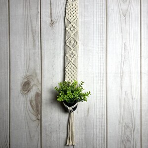 May include: Cream-colored macrame plant hanger with a wooden ring at the top. The hanger features a diamond pattern and holds a small black pot with green foliage. The design includes a long tassel at the bottom, perfect for indoor decor.